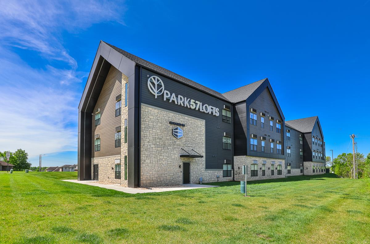 Three-quarter view across the lawn: gabled end with the PARK57LOFTS sign and leaf mark, long wing beyond, blue sky