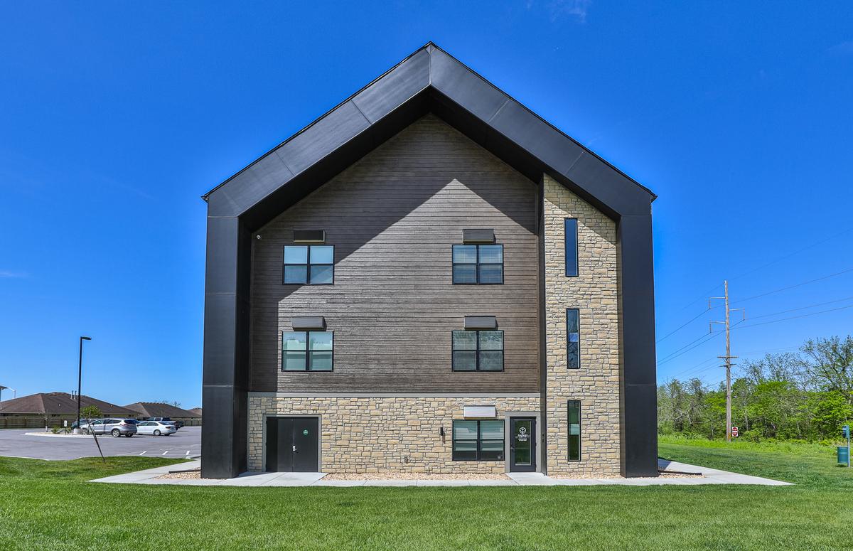 End elevation of the gabled building: dark metal roof and siding, wood-look upper cladding, stone base, blue sky